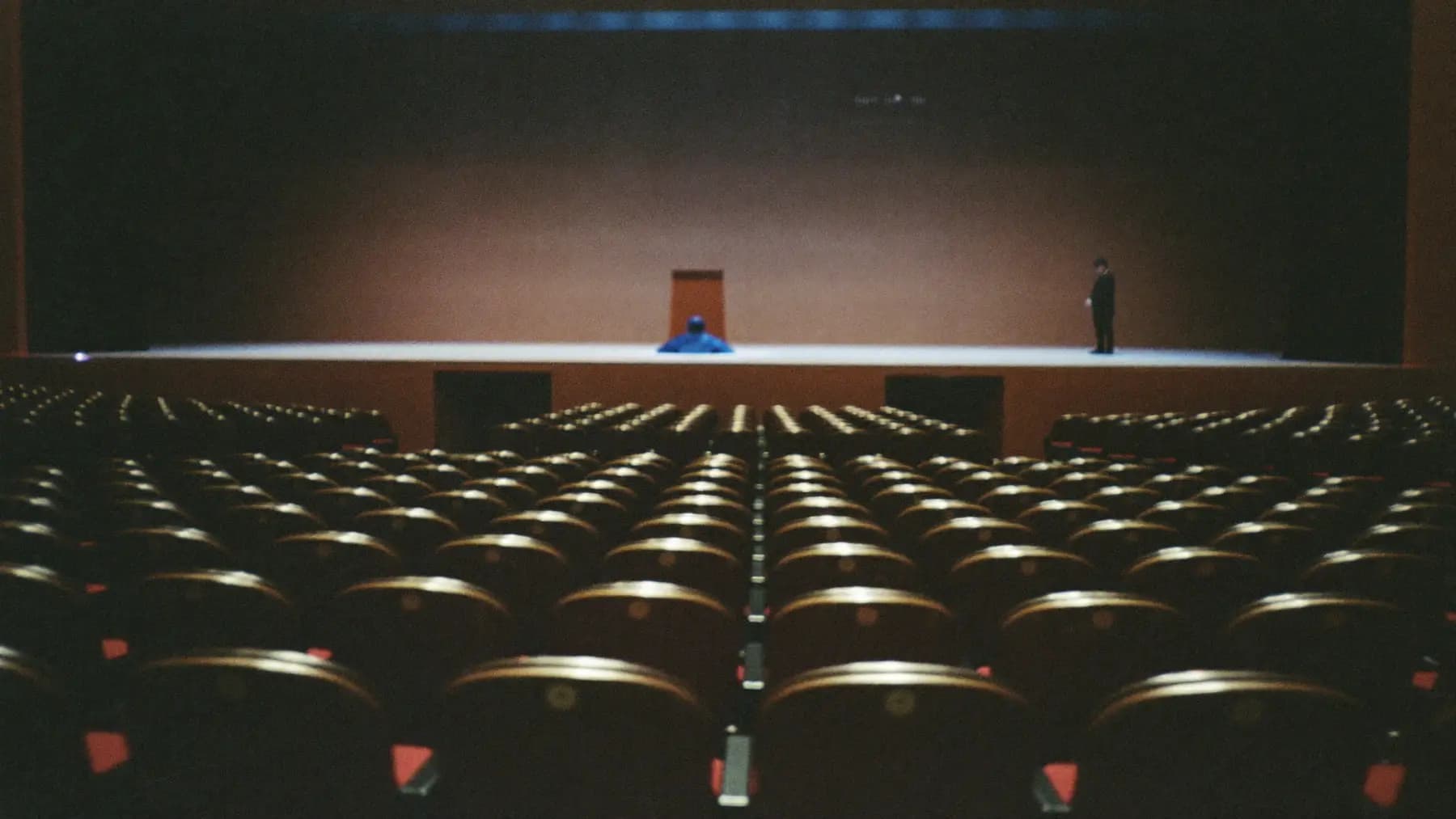 An empty conference auditorium during rehearsal, seats in the foreground, a lit but empty stage in the distance.