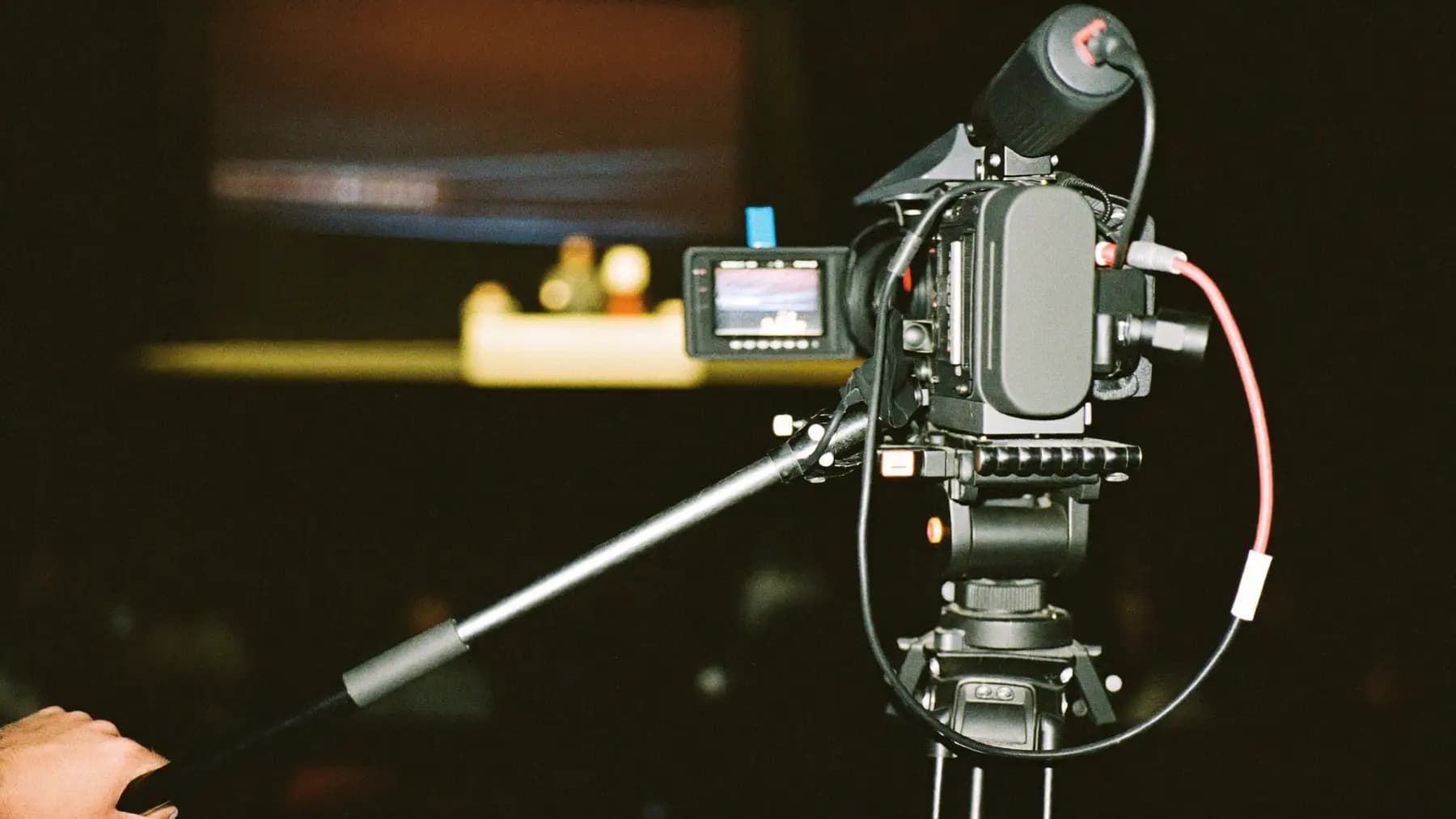 A broadcast camera on a tripod at the back of a darkened conference hall, the operator's hand on the pan bar.