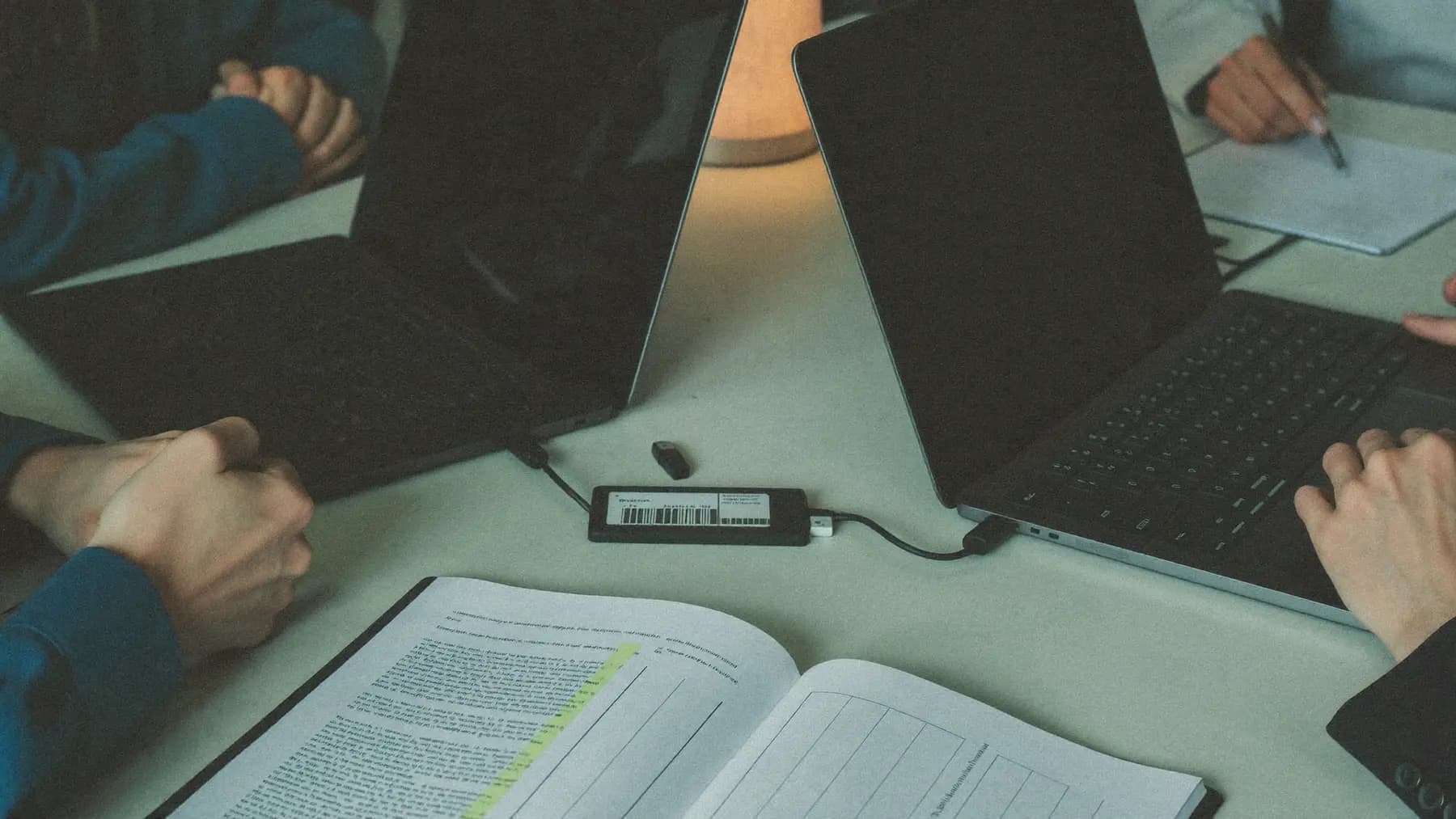 A handover across a meeting table: two laptops facing each other, an open runbook with a section highlighted, drives and an access card between them.