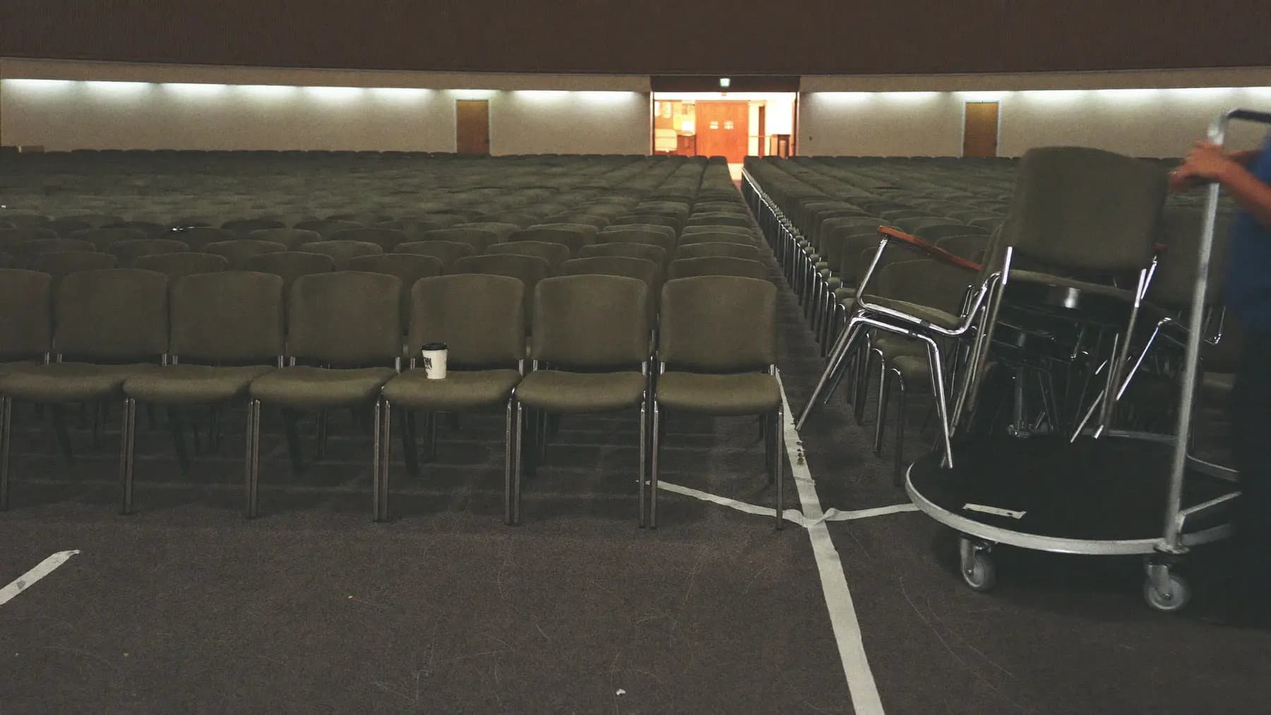 A conference hall after the audience has gone: chairs out of alignment and part-stacked on a trolley, tape being lifted from the carpet.