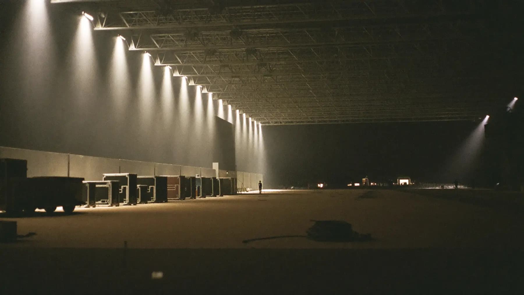 A large convention hall being built the night before an event: half-rigged truss overhead, flight cases lined along one wall, work-lights pooling in a dark empty space.